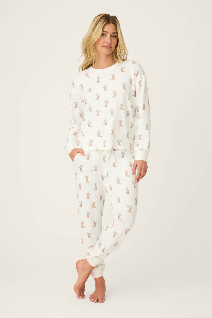 A person stands barefoot against a plain background, wearing the P.J. Salvage Western Blooms Boot Bouquet Lounge Set: a matching white pajama set with a small colorful pattern, long-sleeve top, full-length cuffed pants, and hands in the pants pockets.
