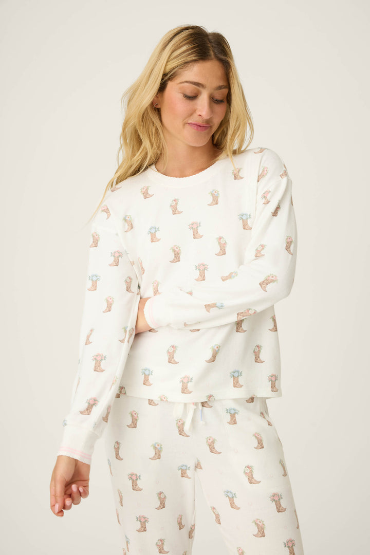 A woman with long blonde hair wears the P.J. Salvage Western Blooms Boot Bouquet Lounge Set, featuring a repeating animal print. She is looking down and smiling slightly with one arm crossed, against a plain light background.