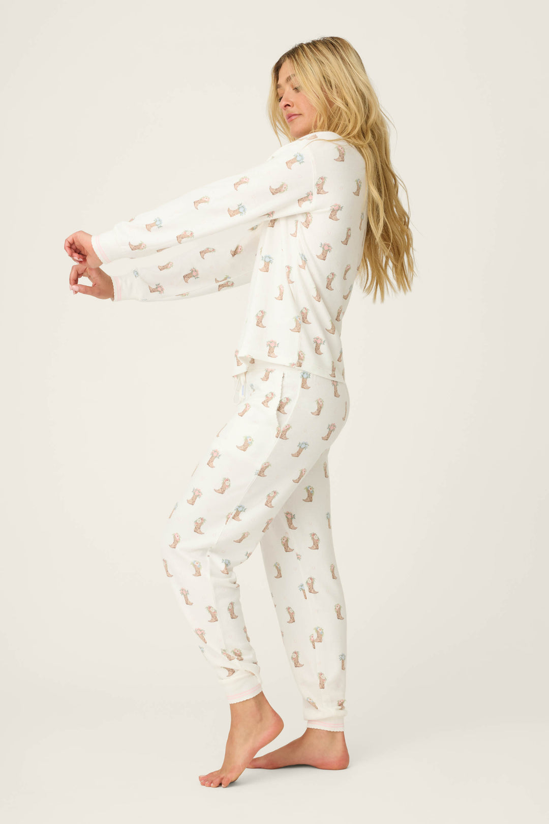 A woman with long blonde hair stands barefoot on a white background, facing sideways with arms extended, wearing the Western Blooms Boot Bouquet Lounge Set by P.J. Salvage featuring a small dog print pattern.