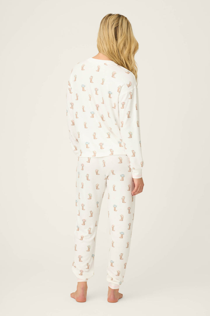 A person with long blonde hair stands barefoot, facing away, wearing the P.J. Salvage Western Blooms Boot Bouquet Lounge Set—white pajamas with a colorful animal pattern on a plain background.