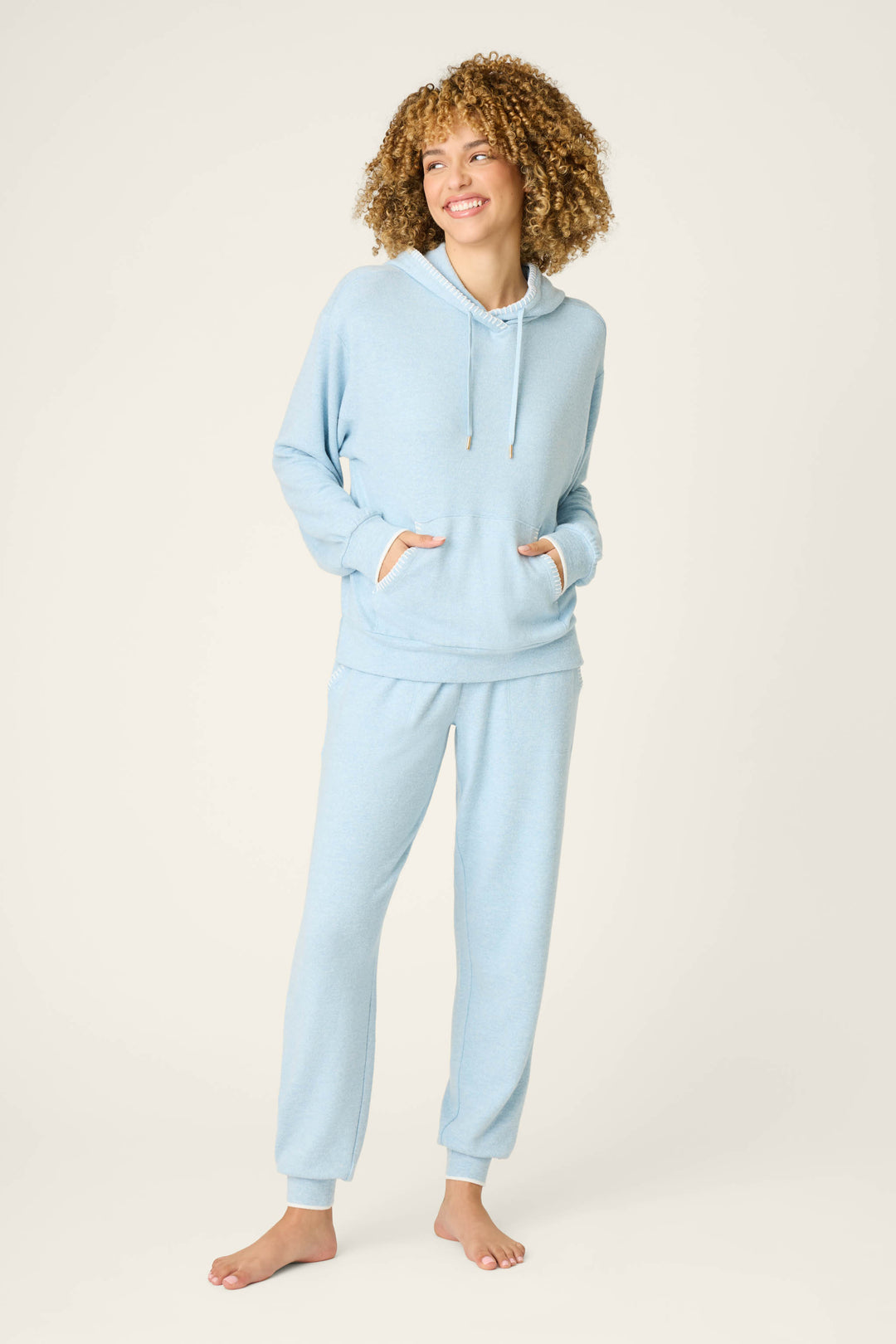 A person stands barefoot against a plain background, smiling and wearing the P.J. Salvage Western Blooms Dream Blue Lounge Set, which features a light blue hooded sweatshirt with front pockets and matching sweatpants.
