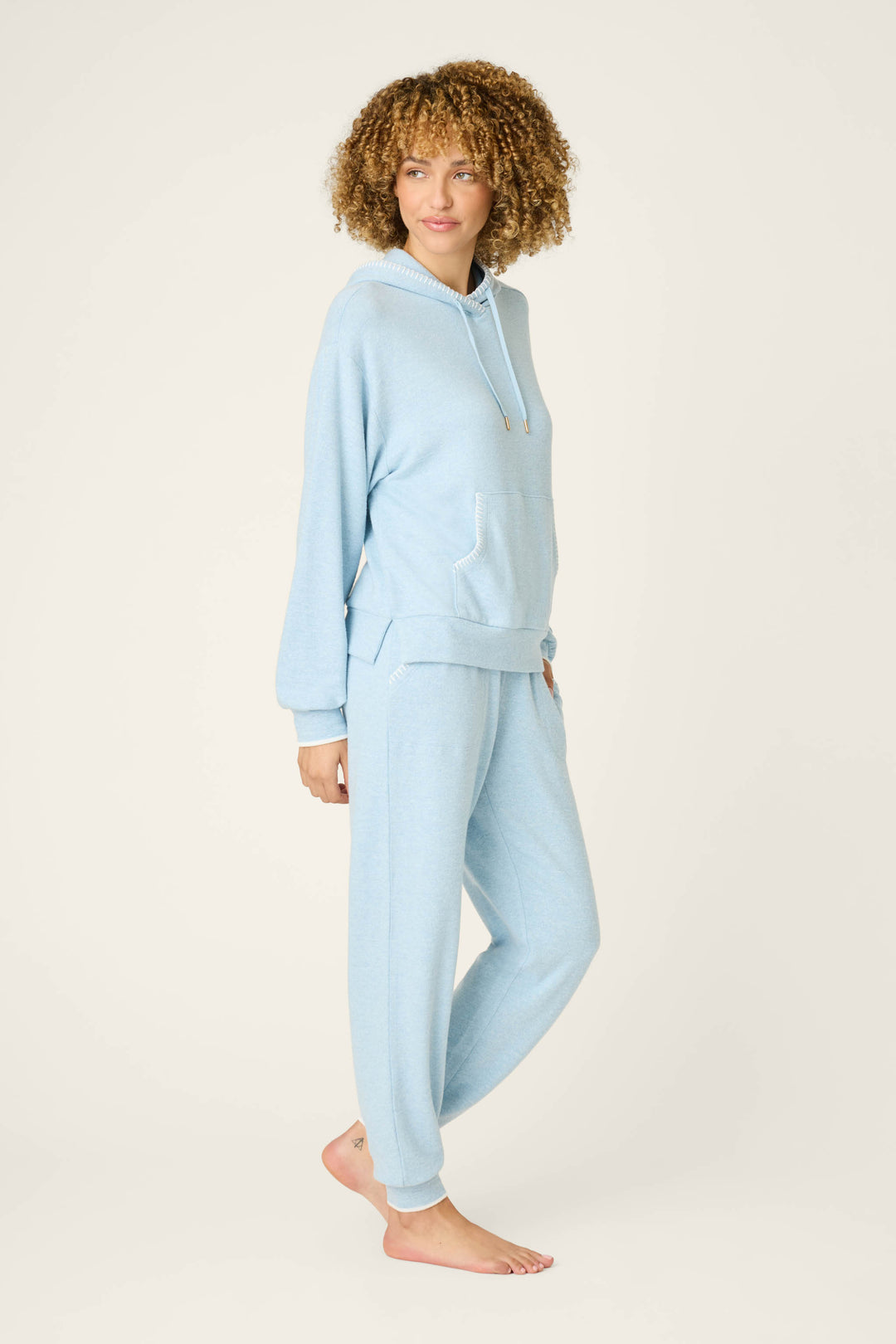 A person with curly hair wears the P.J. Salvage Western Blooms Dream Blue Lounge Set, featuring a light blue hooded sweatshirt and matching jogger pants, standing barefoot on a plain, light background looking slightly to the side.