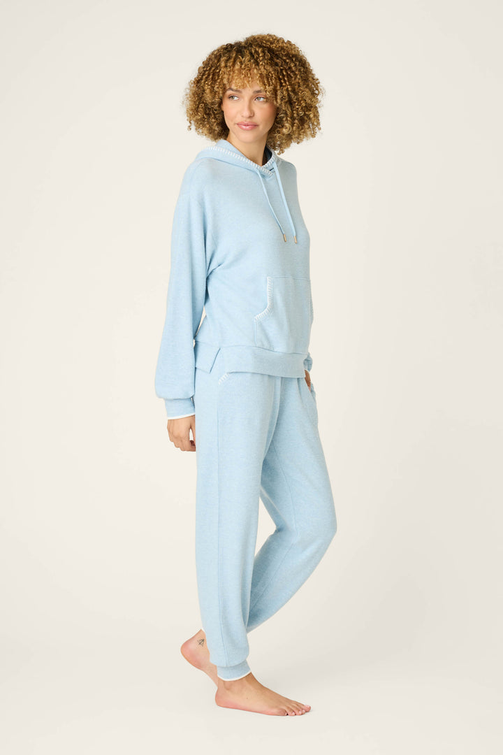 A person with curly hair wears the P.J. Salvage Western Blooms Dream Blue Lounge Set, featuring a light blue hooded sweatshirt and matching jogger pants, standing barefoot on a plain, light background looking slightly to the side.