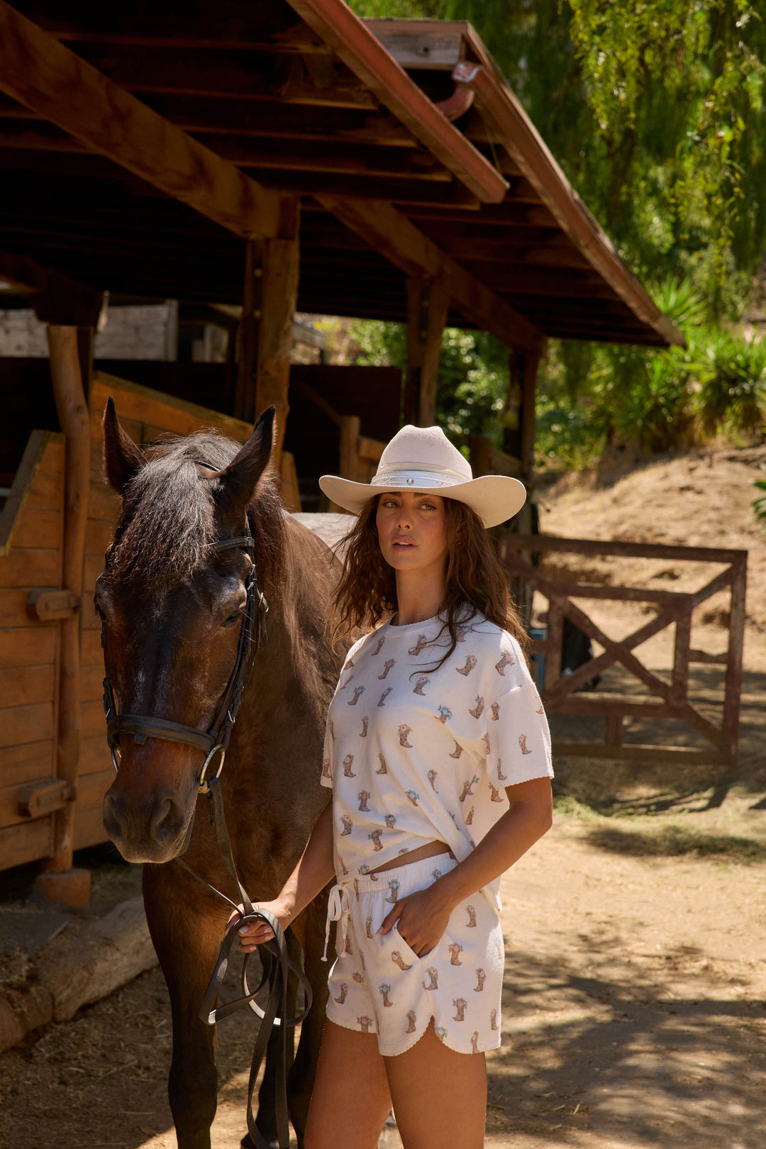 A woman outdoors stands beside a dark brown horse, holding its reins. She wears the P.J. Salvage Western Blooms Boot Bouquet Shorts with a matching white hat and shirt; a wooden stable and trees are in the background.