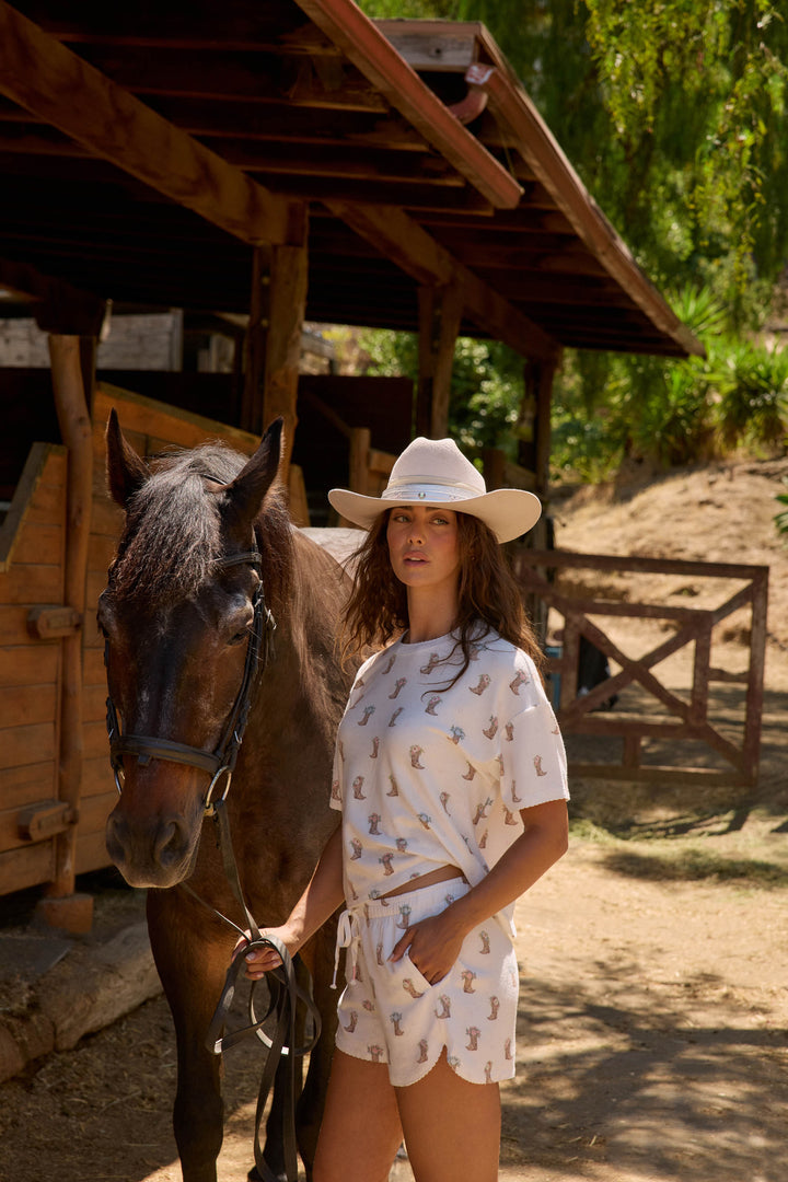 A woman outdoors stands beside a dark brown horse, holding its reins. She wears the P.J. Salvage Western Blooms Boot Bouquet Shorts with a matching white hat and shirt; a wooden stable and trees are in the background.