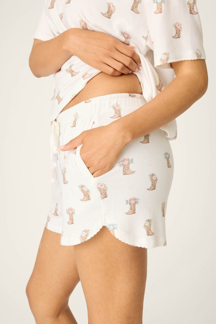 A person models the P.J. Salvage Western Blooms Boot Bouquet Shorts, lifting the shirt to show the waistband while one hand rests in a pocket; the background is plain and light-colored.