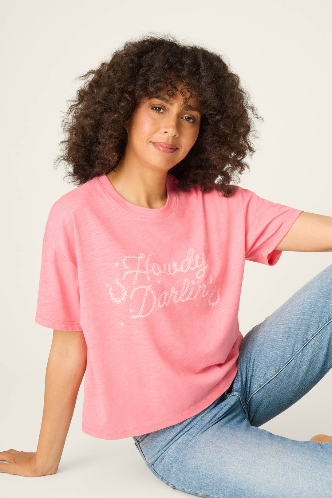 A woman with curly hair wears the P.J. Salvage Western Blooms Howdy Darlin Graphic Print T-Shirt in pink, featuring white script, and light blue jeans as she sits against a light background, smiling slightly with one arm on her knee.