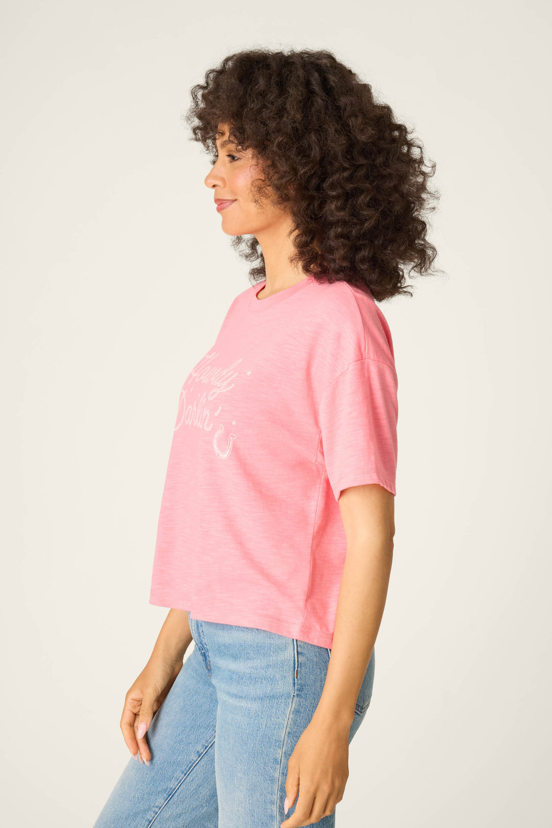 A person with curly hair stands in profile against a plain background, wearing a P.J. Salvage Western Blooms Howdy Darlin graphic print T-shirt and light blue jeans.