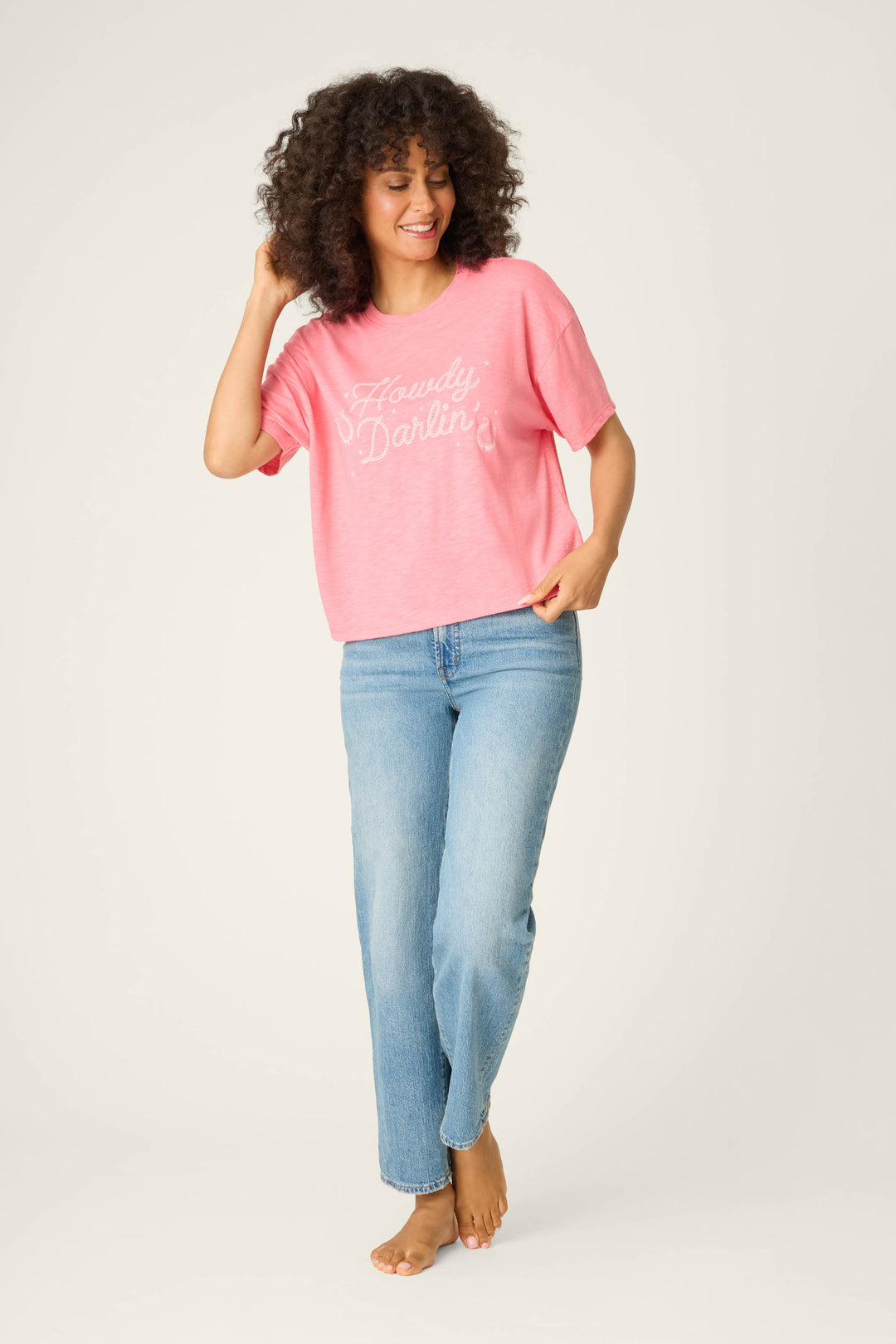 A curly-haired person wears the P.J. Salvage Western Blooms Howdy Darlin Graphic Print T-Shirt in pink with white text, paired with light blue jeans, standing barefoot against a plain light background.