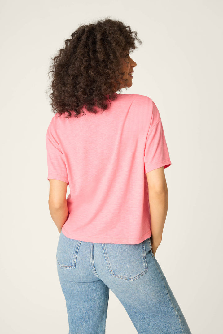 A person with curly hair is seen from the back wearing the P.J. Salvage Western Blooms Howdy Darlin Graphic Print T-Shirt in pink with light blue jeans, standing against a plain, light-colored background.