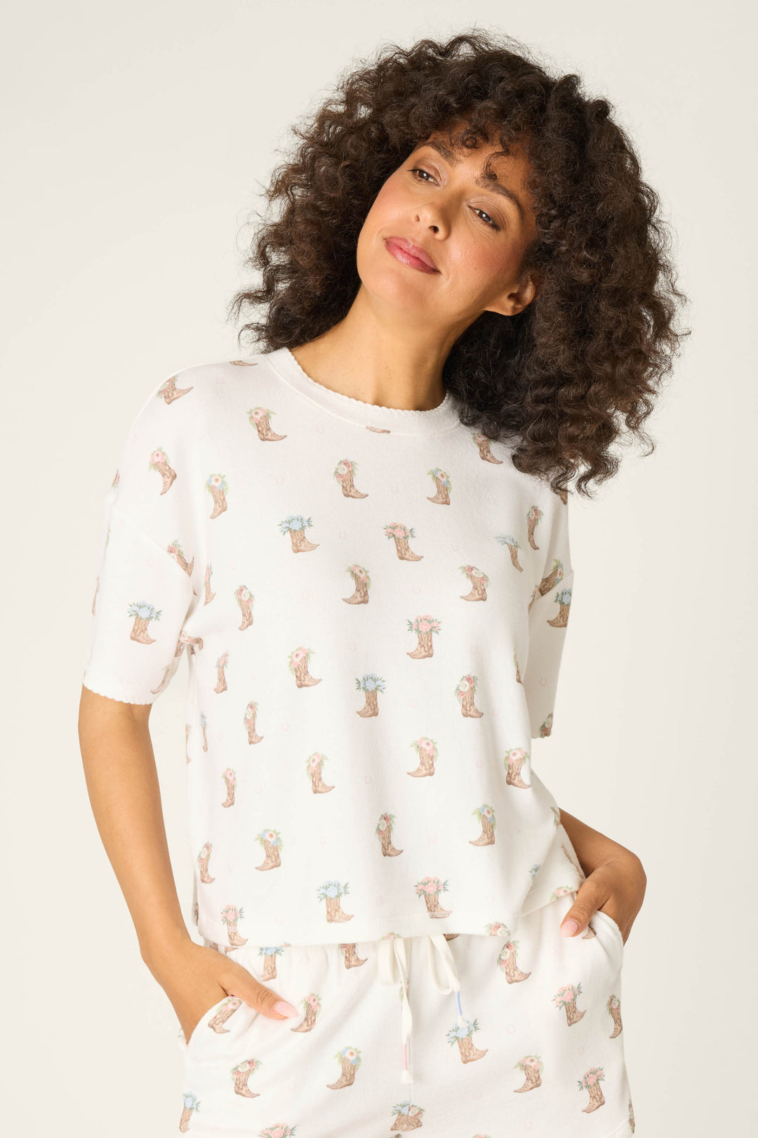 A person with curly hair wears the P.J. Salvage Western Blooms Boot Bouquet Short Sleeve Top—an illustrated white pajama T-shirt with a matching pants set—standing hands in pockets against a plain light background.