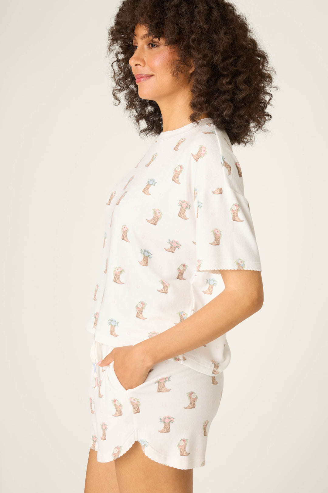 A woman with curly hair stands sideways, wearing the P.J. Salvage Western Blooms Boot Bouquet Short Sleeve Top and matching shorts with pockets, both featuring a repeated illustrated animal pattern. The background is plain light gray.