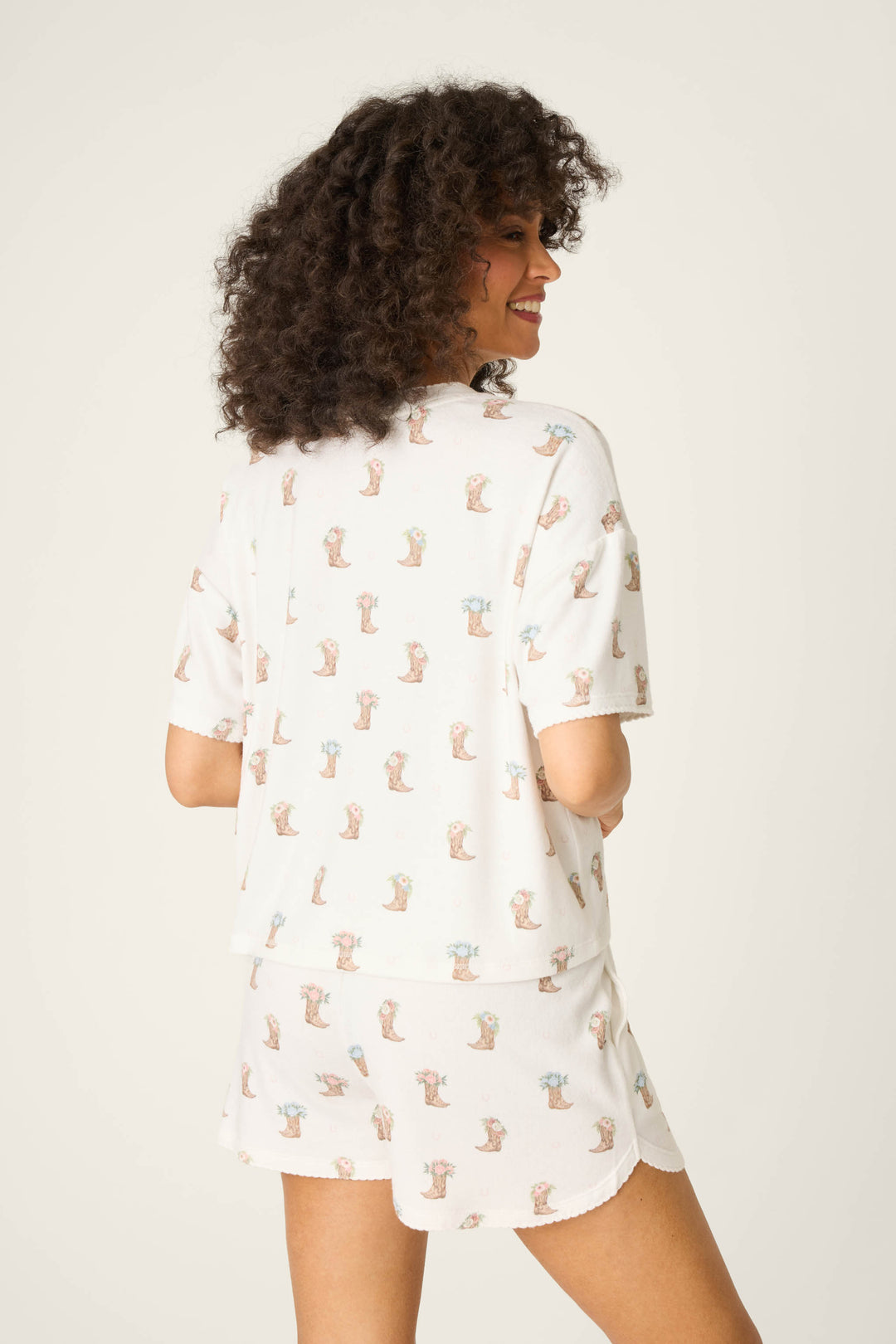 A person with curly hair stands facing away, wearing P.J. Salvage's Western Blooms Boot Bouquet Short Sleeve Top in a soft peachy jersey fabric, which pops against the plain, light background.