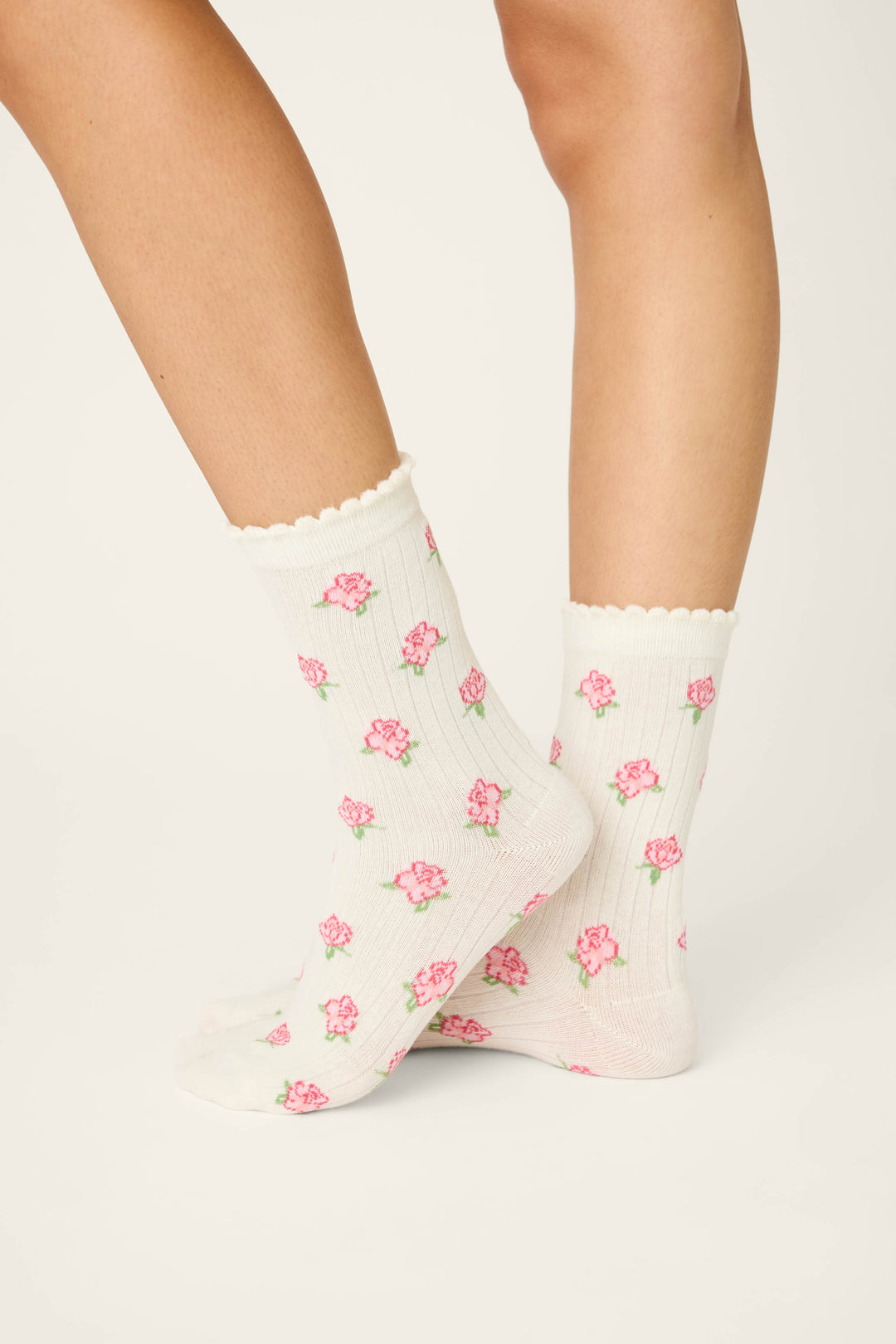 A pair of legs wearing P.J. Salvage Western Blooms Roses Ribbed Crew Socks, featuring an ivory base with scalloped edges and a delicate pink floral design, are crossed over each other against a plain background.
