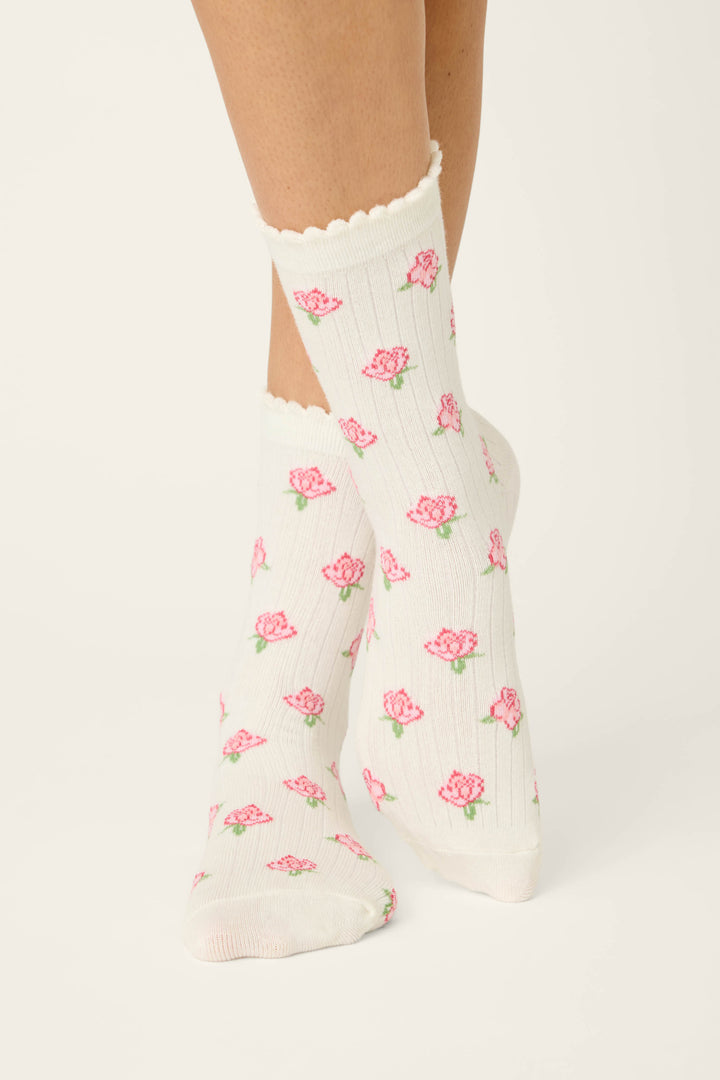 A person models the P.J. Salvage Western Blooms Roses Ribbed Crew Socks, featuring a pink floral pattern and scalloped edge in ivory, standing with one foot crossed before the other against a plain background.