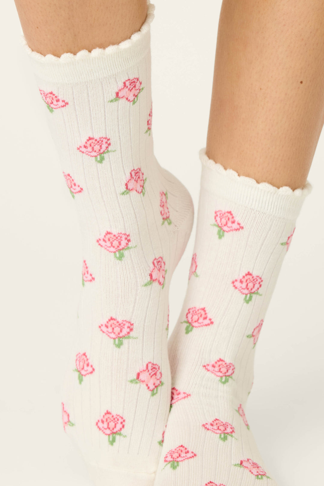 A close-up of two feet in P.J. Salvage Western Blooms Roses Ribbed Crew Socks, featuring an ivory ribbed texture, scalloped edge, and a delicate pattern of pink roses with green leaves.