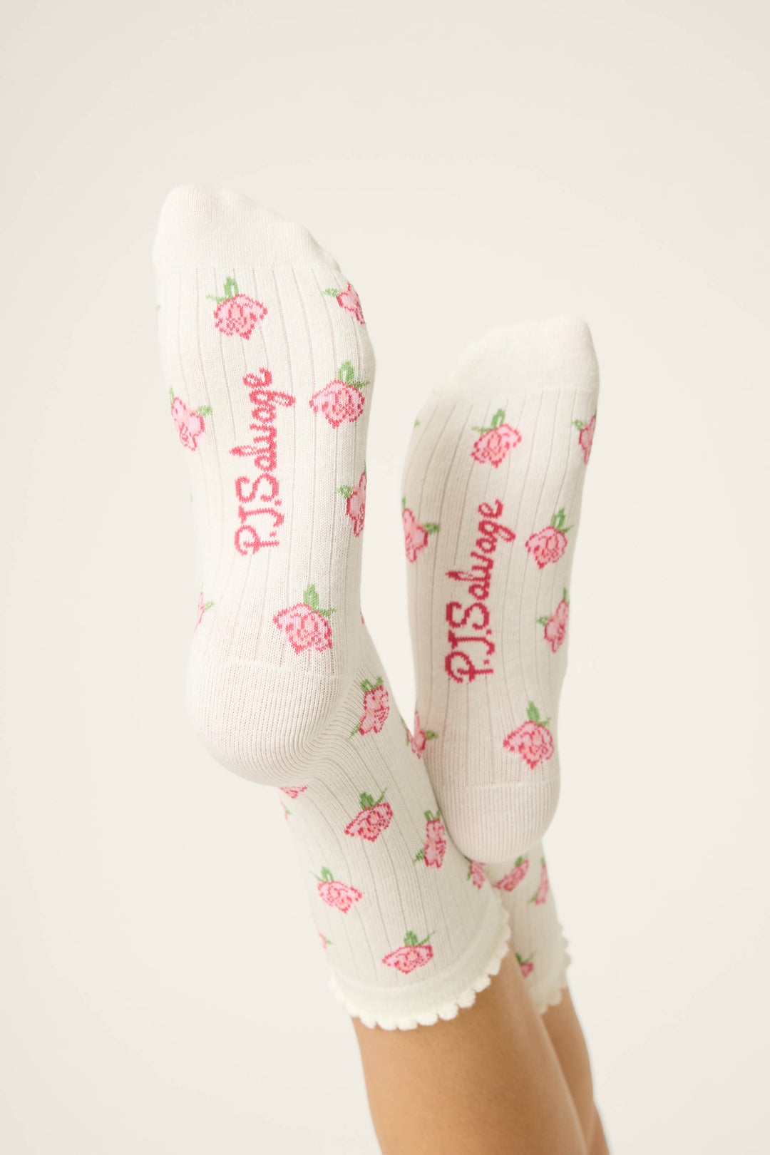A person’s feet in Western Blooms Roses Ribbed Crew Socks by P.J. Salvage, featuring an ivory base with a pink rose motif and the brand’s name in pink on the soles. The scalloped-edged socks are raised against a plain background.