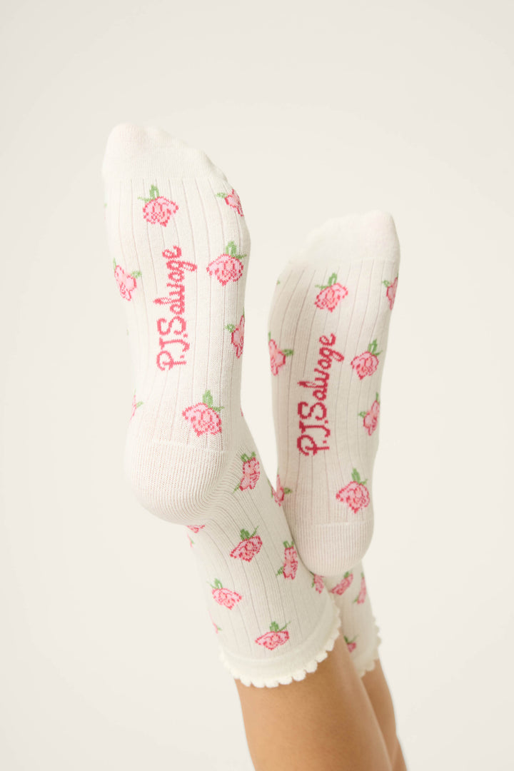 A person’s feet in Western Blooms Roses Ribbed Crew Socks by P.J. Salvage, featuring an ivory base with a pink rose motif and the brand’s name in pink on the soles. The scalloped-edged socks are raised against a plain background.