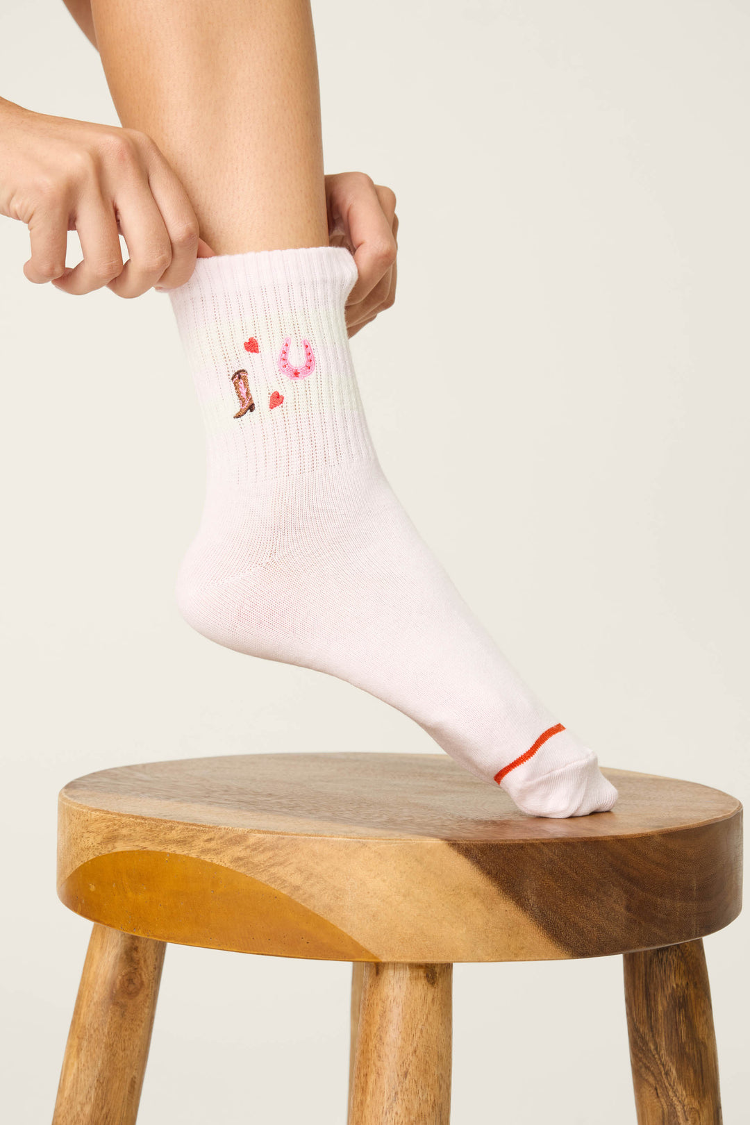 A person wearing P.J. Salvage Western Blooms Crew Socks in ballet-pink, featuring small embroidered icons like a horseshoe and red heart, adjusts their sock while resting a foot on a wooden stool.