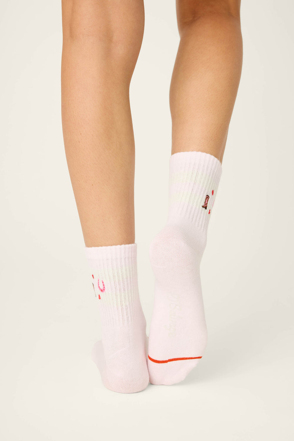 A person faces away from the camera, displaying lower legs and feet in P.J. Salvage Western Blooms Crew Socks—white ribbed knit crew socks with subtle red and green accents—set against a plain light background.
