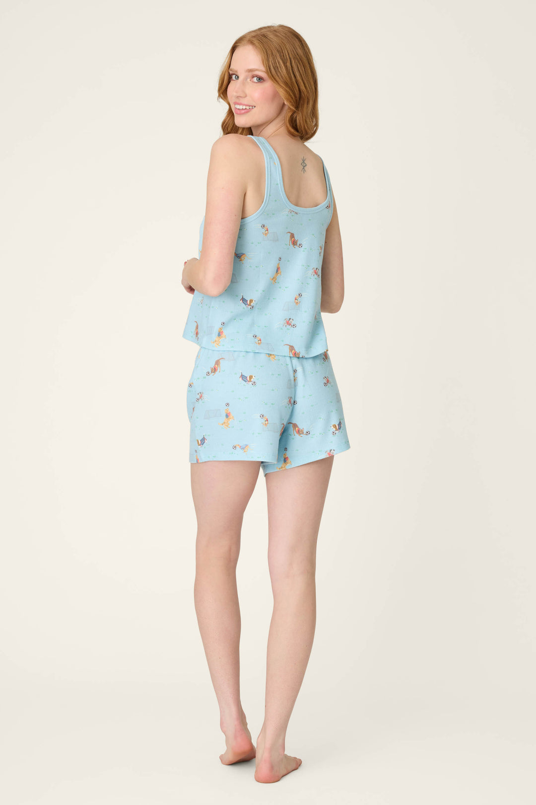 A woman with long red hair stands barefoot, facing away but looking over her shoulder, against a plain off-white background.