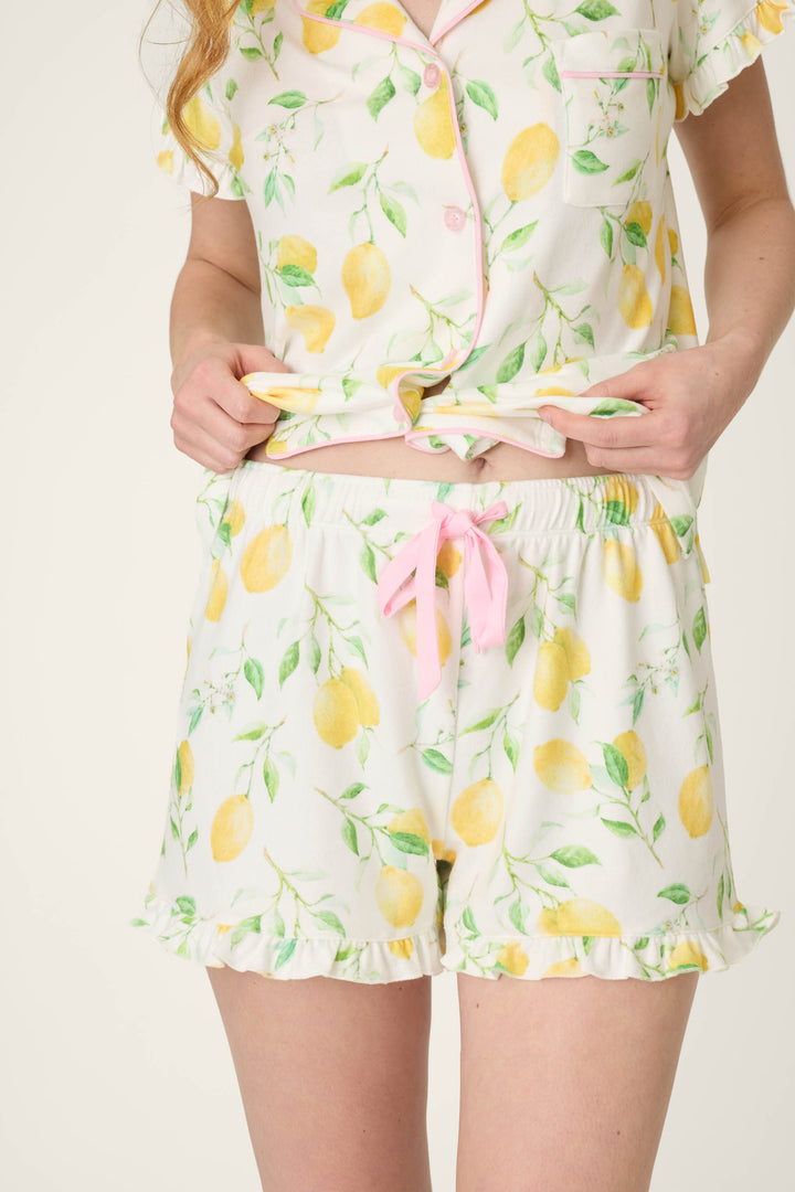 White fabric with yellow lemon and green leaf print, accented by pink buttons, pink ribbon, and ruffled hems.