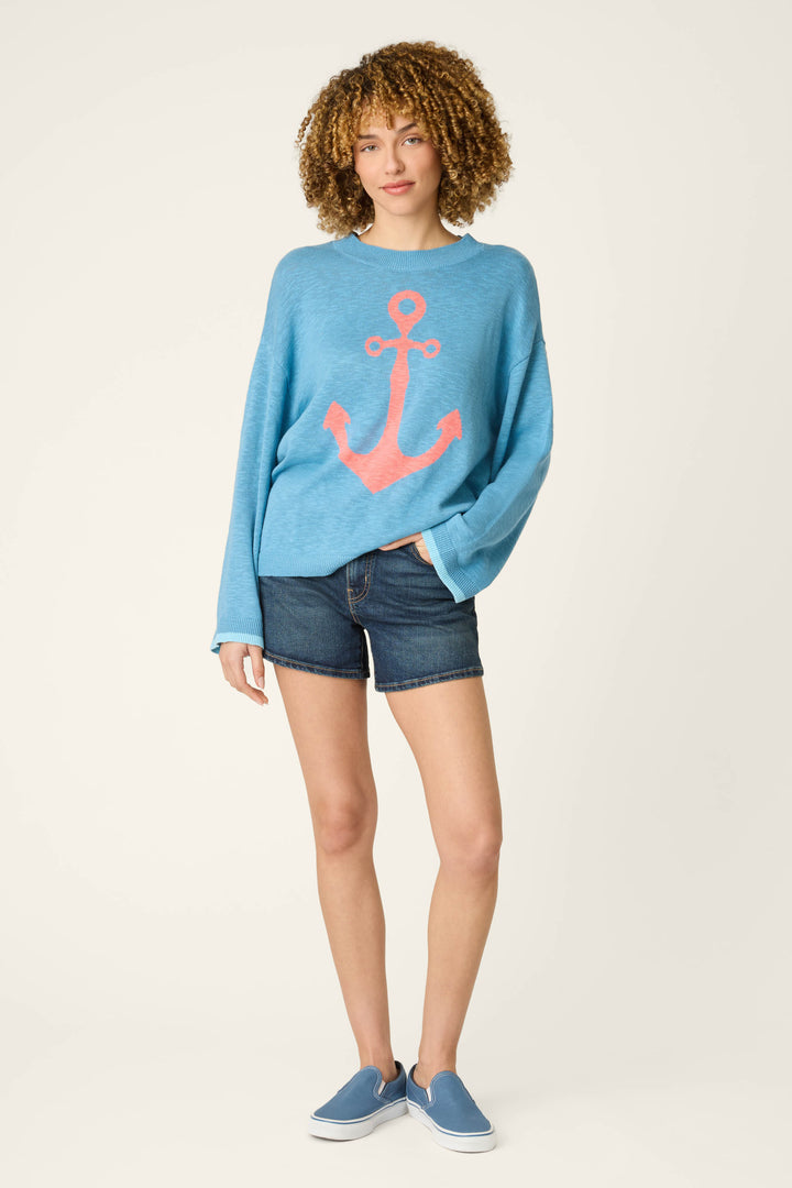 A person with curly hair wears a sweater with a large pink anchor graphic, dark denim shorts, and blue slip-on shoes on light background.