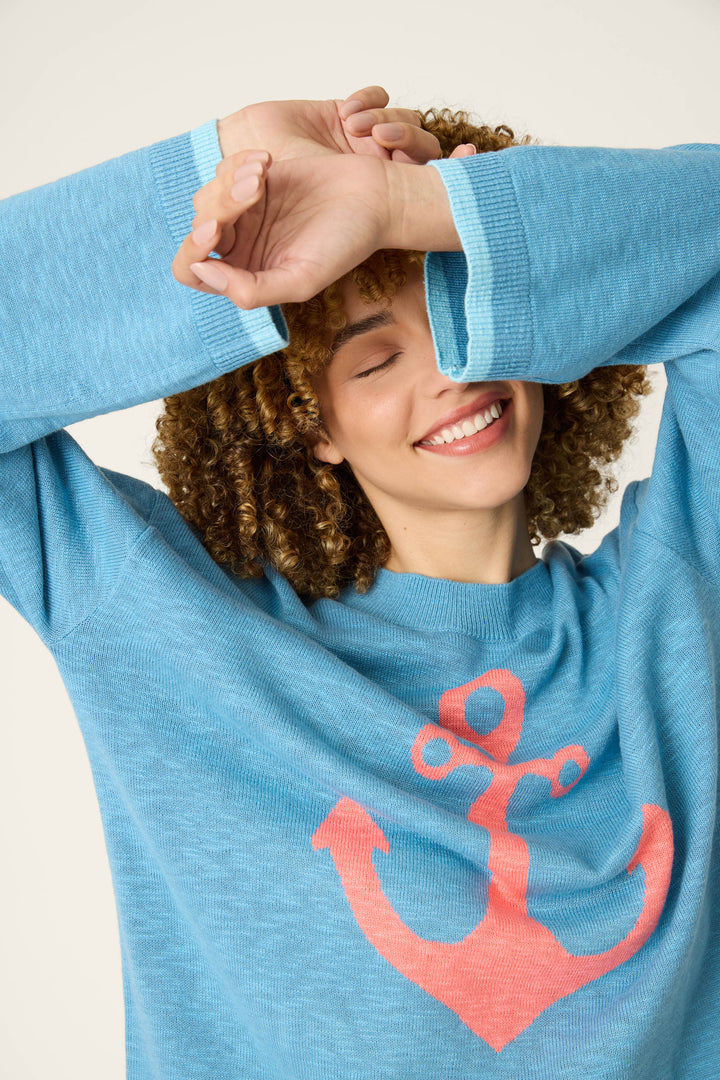 A person with curly hair smiles with eyes closed, raising one arm to partially cover their face while wearing a sweater with a large pink anchor on the front.