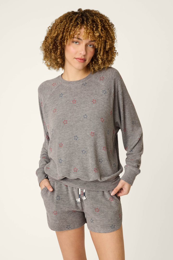 A person with curly hair wears the P.J. Salvage Red, White, & Woof Star Print Long Sleeve Top—a relaxed fit gray lounge top with small red and blue stars—paired with matching drawstring shorts, hands in pockets against a plain background.
