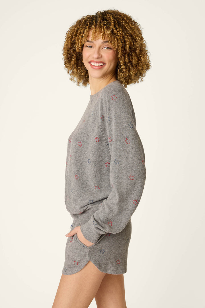 A person with curly hair smiles sideways against a plain background, wearing the P.J. Salvage Red, White, & Woof Star Print Long Sleeve Top—a relaxed fit grey sweatshirt with small stars.