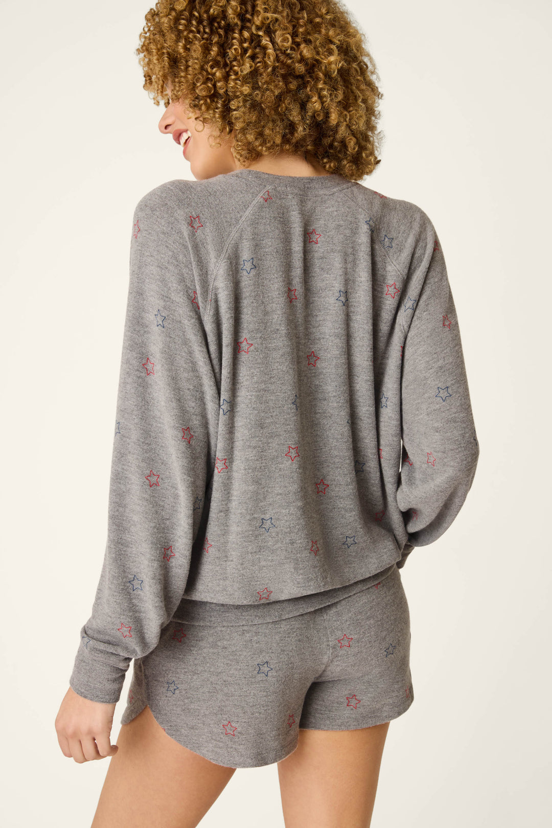 A woman with curly hair smiles while wearing the P.J. Salvage Red, White, & Woof Star Print Long Sleeve Top, a relaxed fit peachy grey jersey lounge set featuring small red and blue stars.