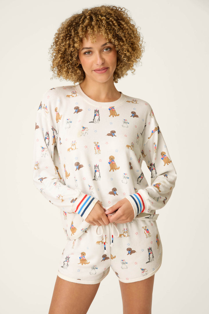 A person with curly hair smiles in the P.J. Salvage Red, White, & Woof Patriotic Pups Long Sleeve Top, featuring playful pup prints and red, blue, and white striped cuffs, set against a light background.