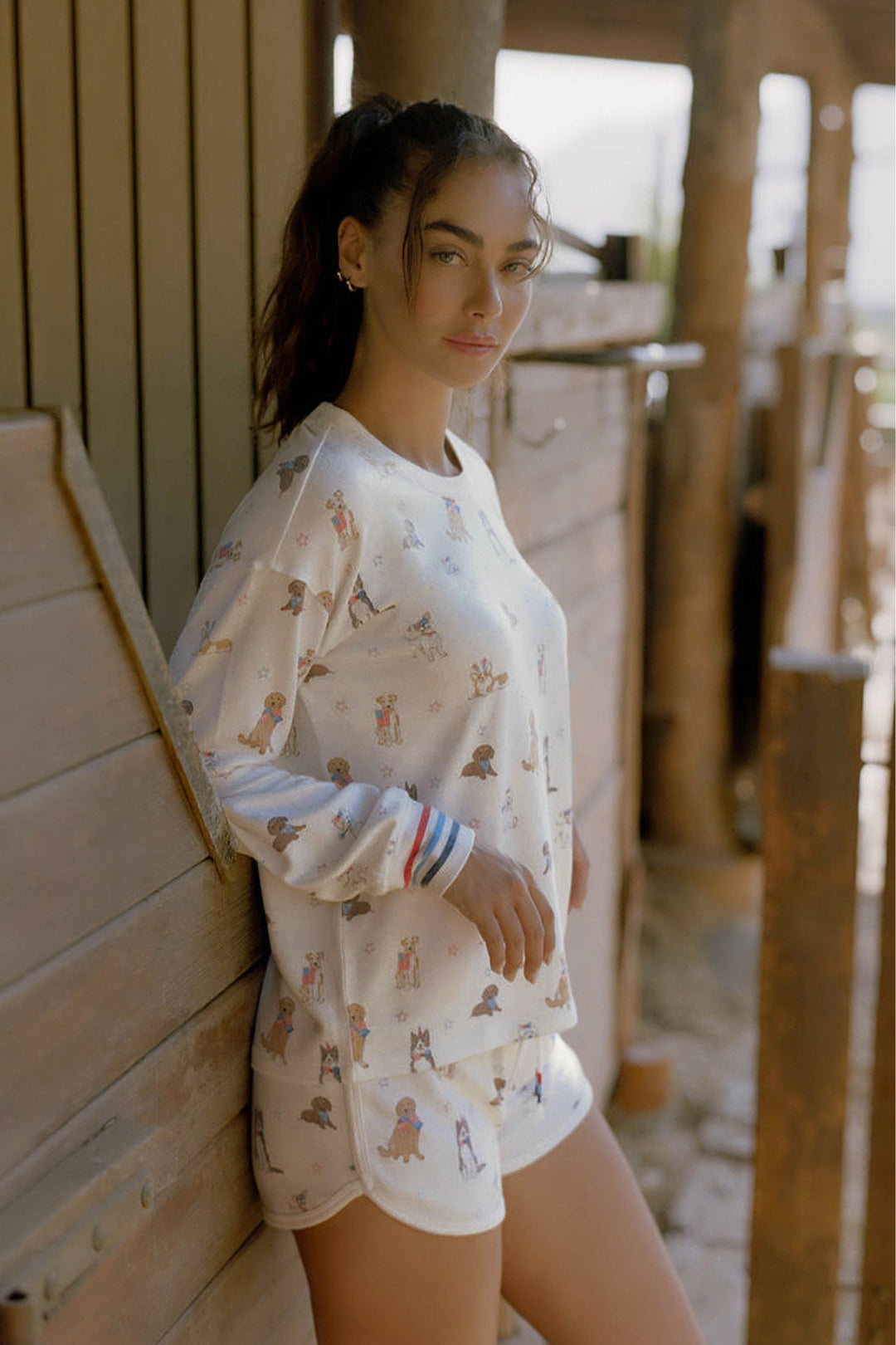 A woman with long dark hair in a ponytail leans against a wooden wall, wearing the P.J. Salvage Red, White, & Woof Patriotic Pups Long Sleeve Top. Sunlight fills the rustic space as she looks at the camera with a relaxed expression.