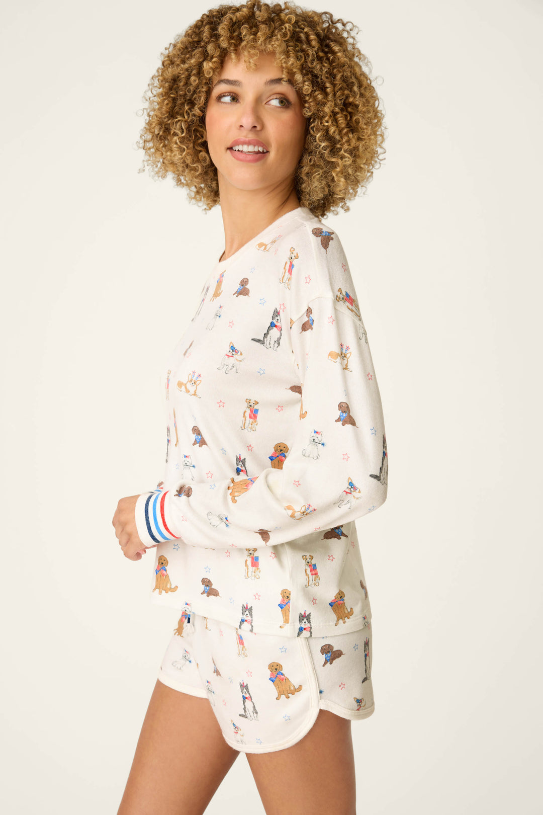 A woman with curly hair smiles while wearing the P.J. Salvage Red, White, & Woof Patriotic Pups Long Sleeve Top—cream-colored with playful pup prints—against a plain light background.