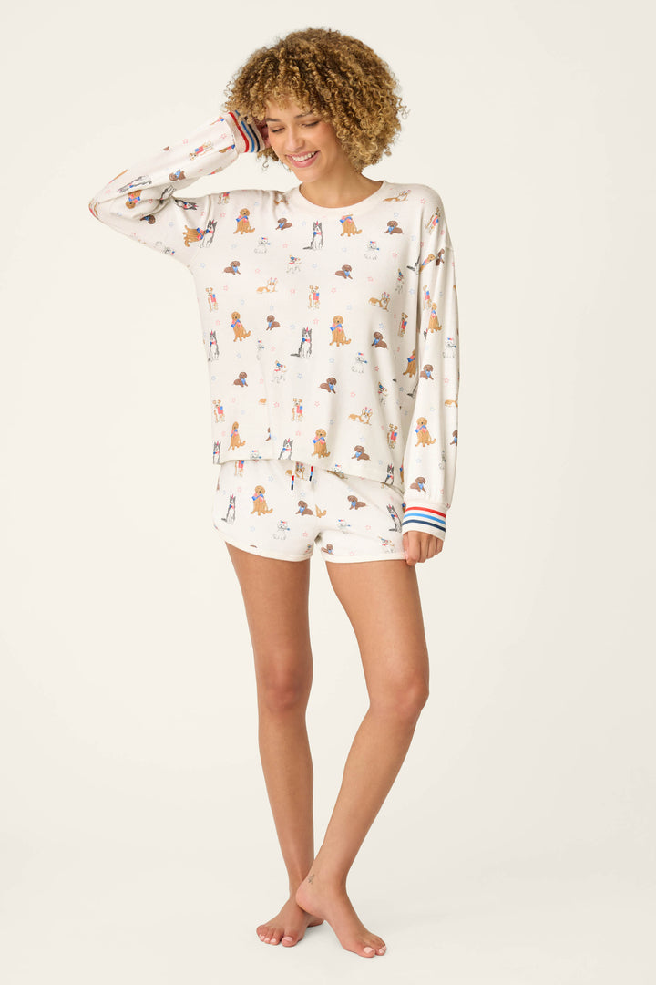 A person with curly hair smiles, wearing the P.J. Salvage Red, White, & Woof Patriotic Pups Long Sleeve Top featuring a playful pup print. They stand barefoot against a plain light background.