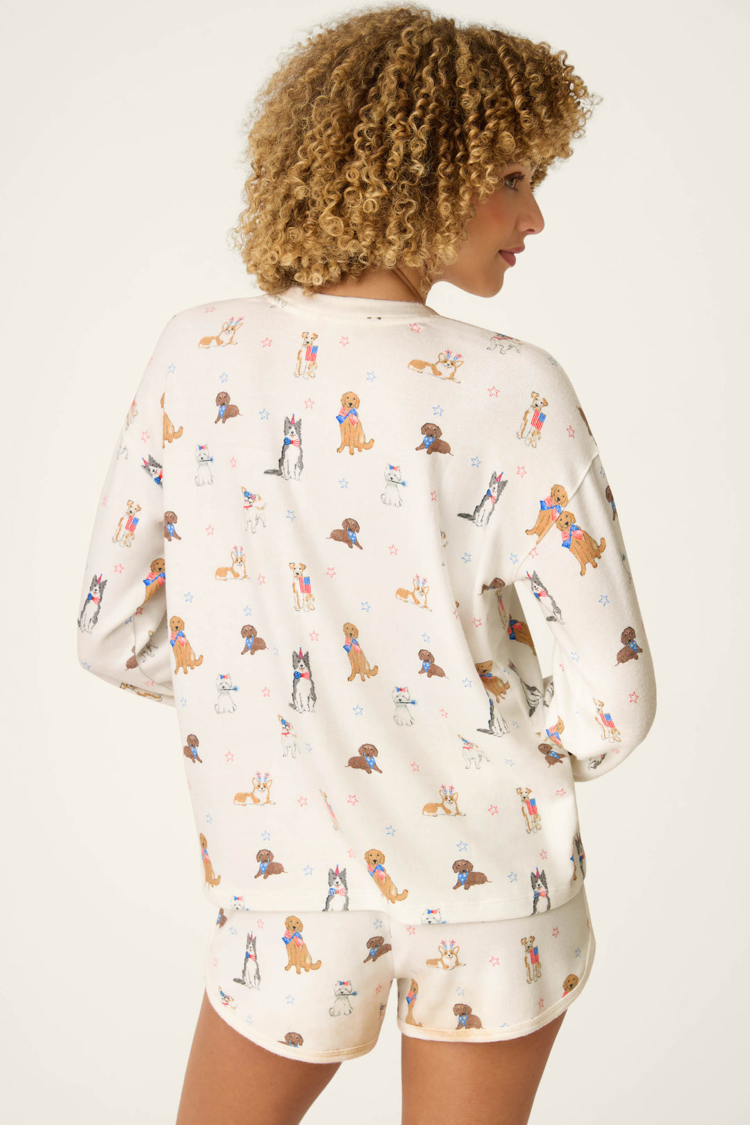 A person with curly hair is seen from the back wearing the P.J. Salvage Red, White, & Woof Patriotic Pups Long Sleeve Top, featuring playful cartoon dogs in red, white, and blue prints.