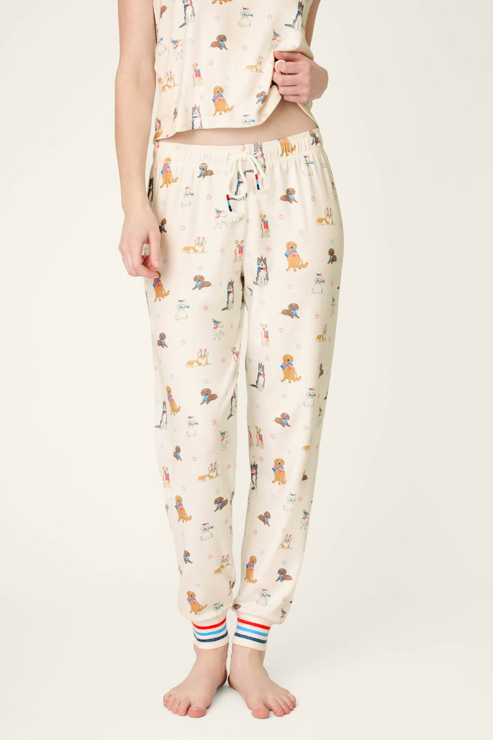 A person stands barefoot in P.J. Salvage Red, White, & Woof Patriotic Pups Banded Pants with pup prints and striped cuffs; the matching pajama top is partially shown against a plain, light background for a cozy loungewear look.