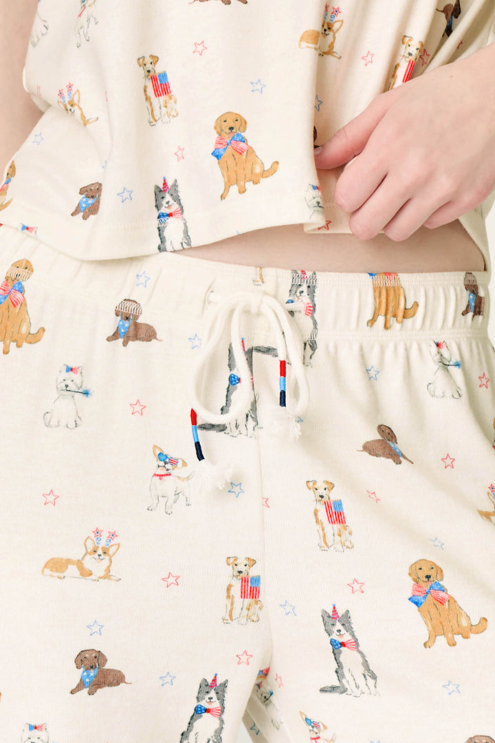 Close-up of a person wearing P.J. Salvage Red, White, & Woof Patriotic Pups Banded Pants in soft peachy jersey, featuring a pup print with dogs in patriotic outfits and red, white, and blue stars; hand rests near the drawstring waistband.