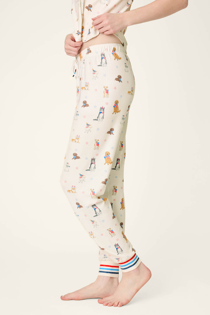 A person in P.J. Salvage Red, White, & Woof Patriotic Pups Banded Pants with multi-colored striped cuffs stands barefoot on a light background. The playful dog print and soft fabric offer cozy comfort for relaxed days at home. Upper body partially visible.