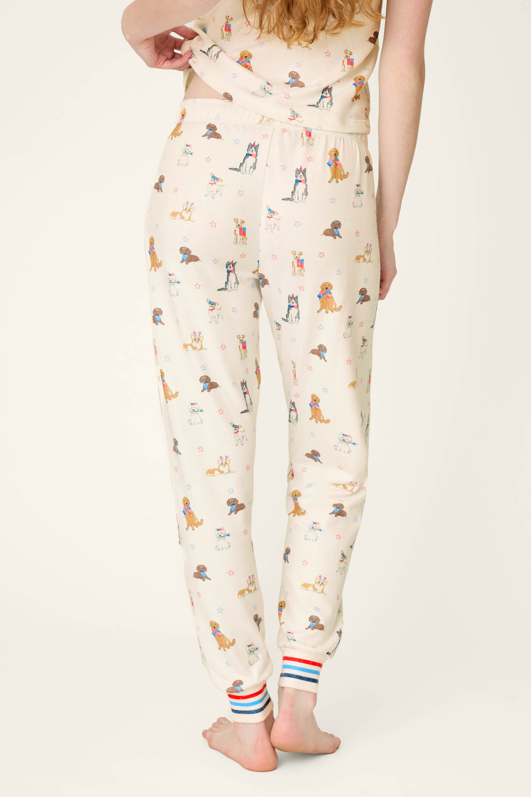 A person stands barefoot, facing away, wearing P.J. Salvage Red, White, & Woof Patriotic Pups Banded Pants—cream-colored with pup print and red, white, and blue striped cuffs.