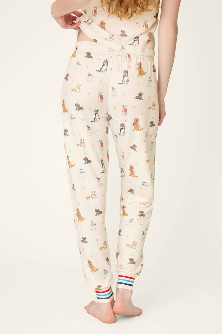 A person stands barefoot, facing away, wearing P.J. Salvage Red, White, & Woof Patriotic Pups Banded Pants—cream-colored with pup print and red, white, and blue striped cuffs.