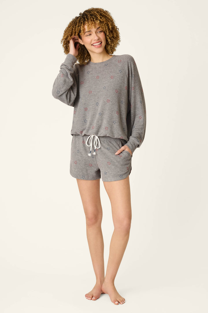 A woman with curly hair smiles while wearing P.J. Salvage’s Red, White, & Woof Star Print Shorts with an elastic waistband and a loose gray long-sleeve top, standing barefoot against a plain background.