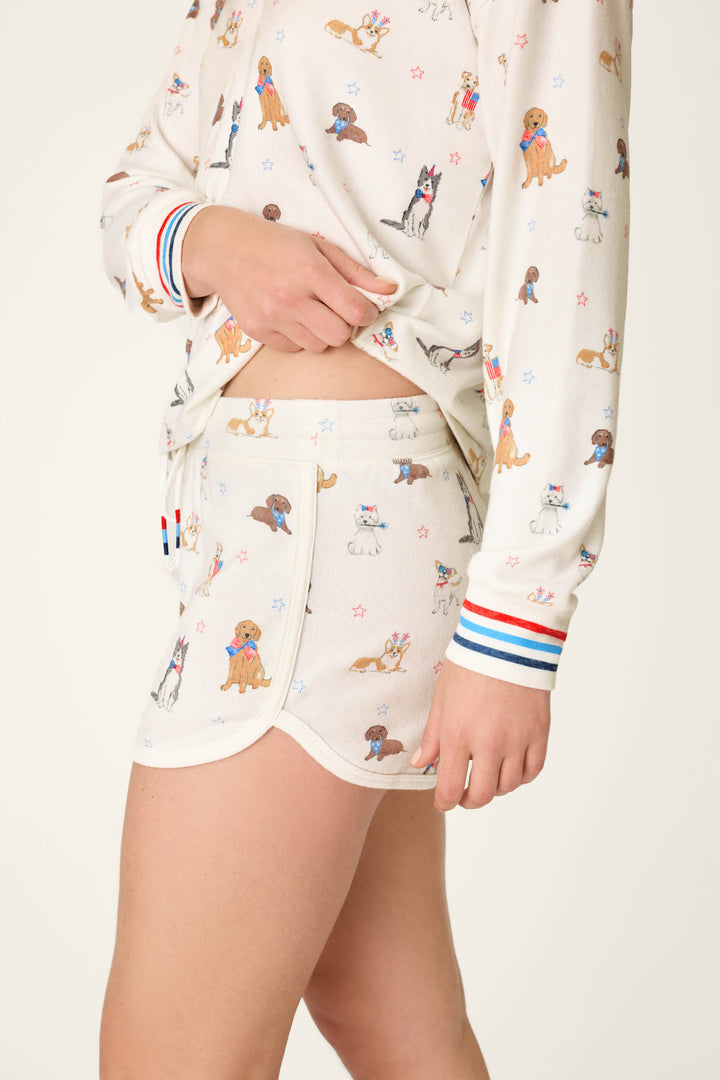 A person wears P.J. Salvage’s Red, White, & Woof Patriotic Pups Shorts with a playful pup and star print, red, white, and blue striped cuffs, and a comfy elastic waistband. Only their lower face and legs are seen.