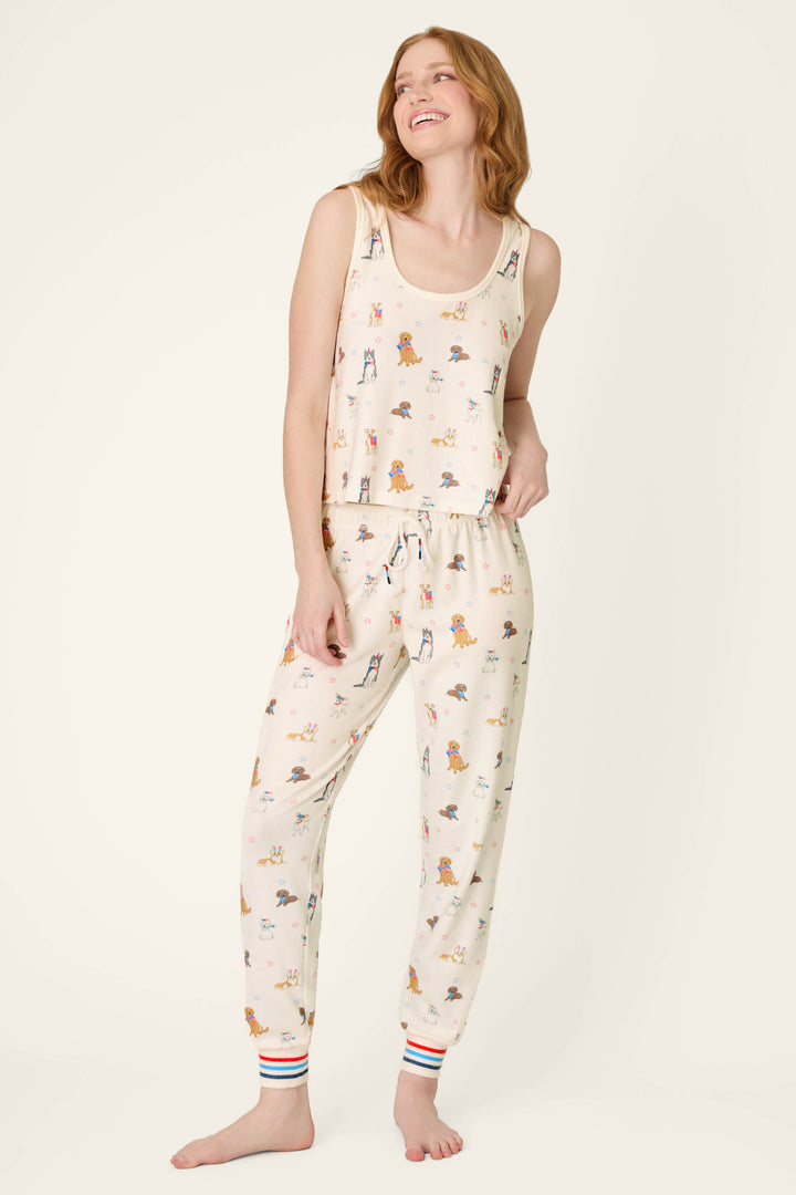 Barefoot woman with wavy light brown hair wears P.J. Salvage's Red, White, & Woof Patriotic Pups Long PJ Set—a cream pajama set with colorful pup print, sleeveless top, and jogger pants—against a light background.