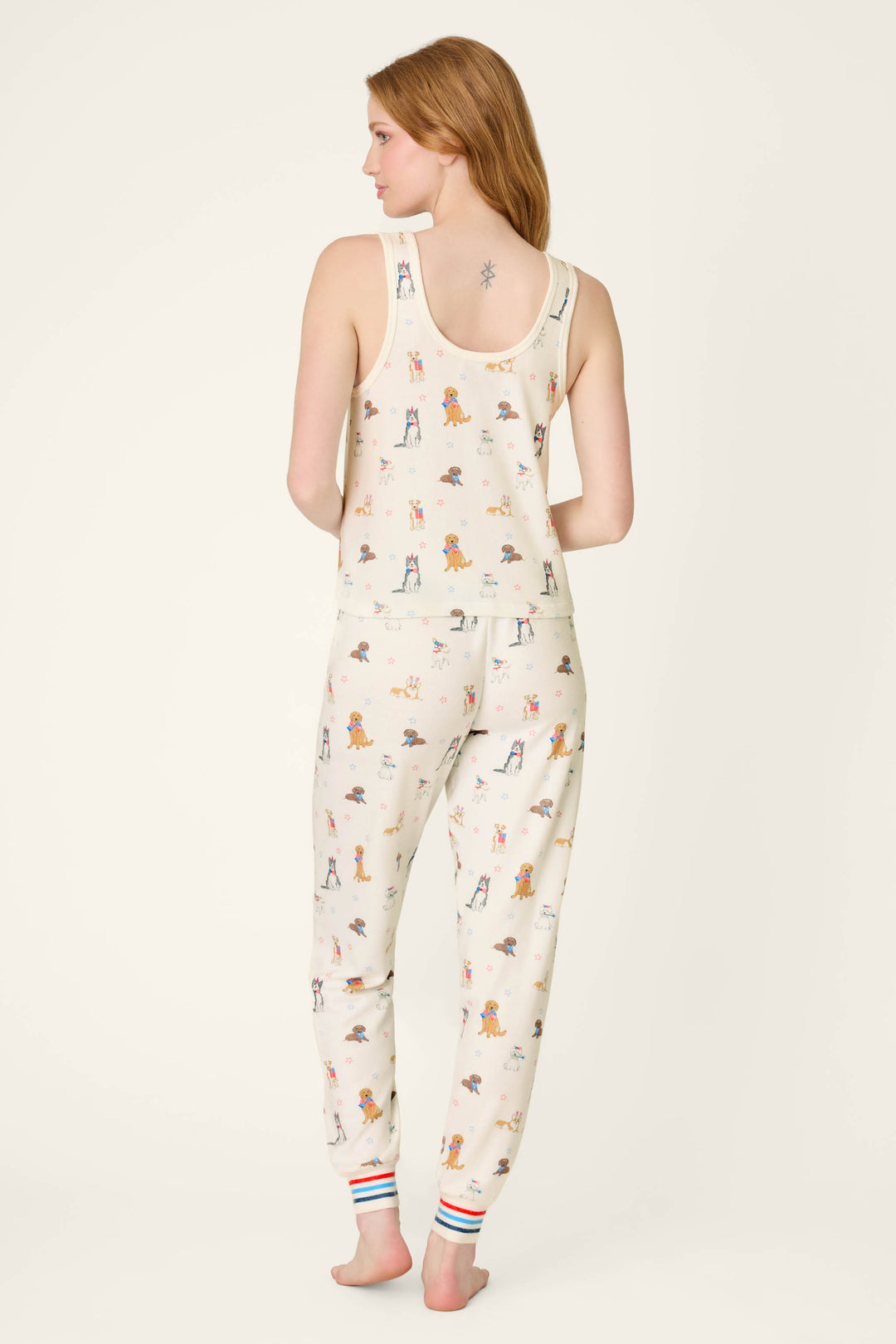 A woman faces away barefoot, wearing the P.J. Salvage Red, White, & Woof Patriotic Pups Long PJ Set—cream-colored with pup prints and striped cuffs. Her long hair is loose, showing a small tattoo on her upper back—ideal summer sleepwear.