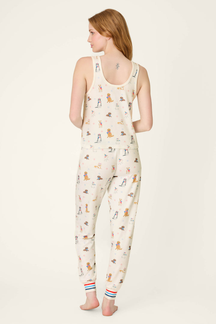 A woman faces away barefoot, wearing the P.J. Salvage Red, White, & Woof Patriotic Pups Long PJ Set—cream-colored with pup prints and striped cuffs. Her long hair is loose, showing a small tattoo on her upper back—ideal summer sleepwear.