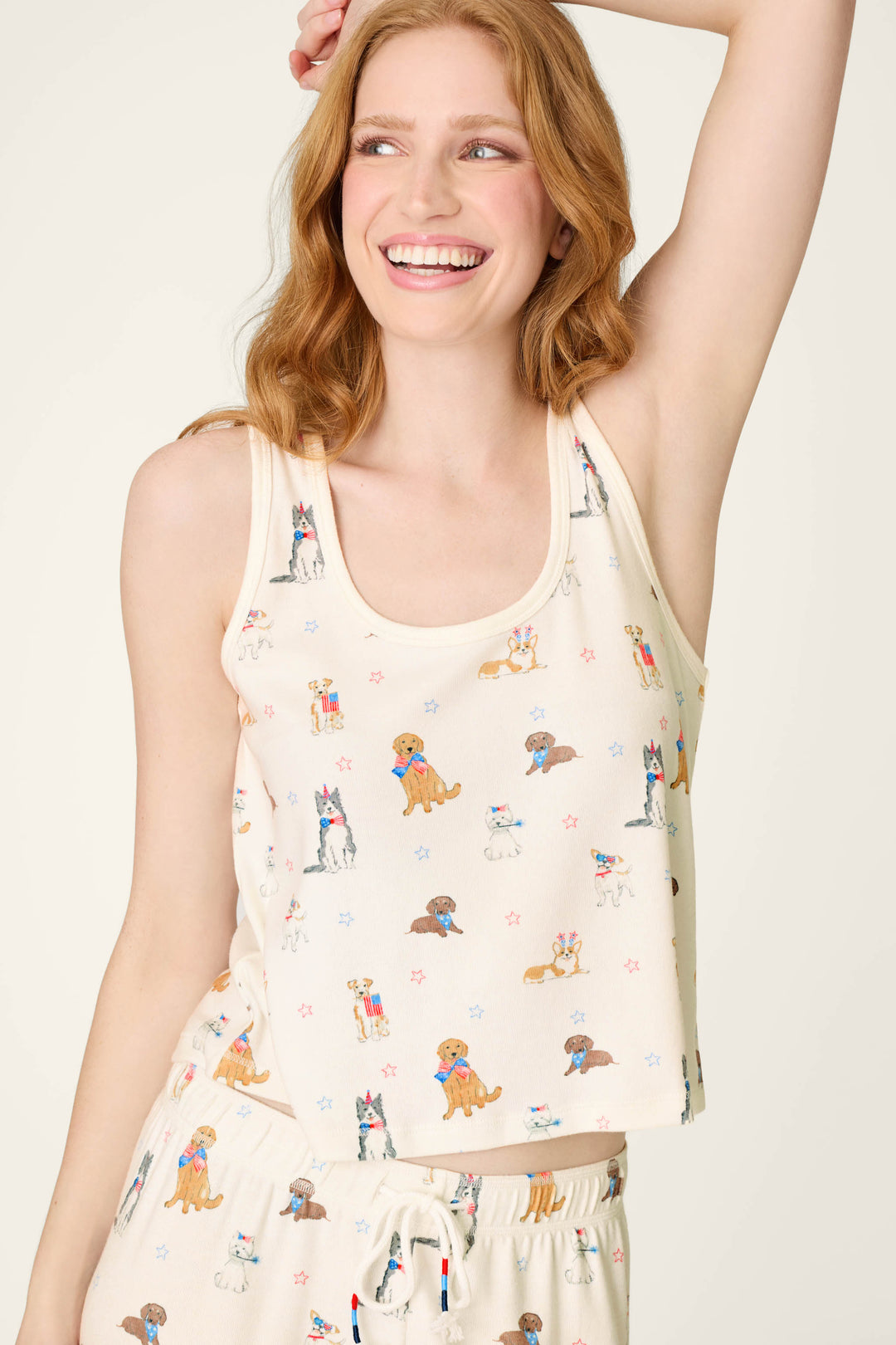 A smiling woman with red hair wears the P.J. Salvage Red, White, & Woof Patriotic Pups Tank Top, a cream scoop neck featuring a playful dog print. She poses with one arm raised, looking to the side against a neutral background.