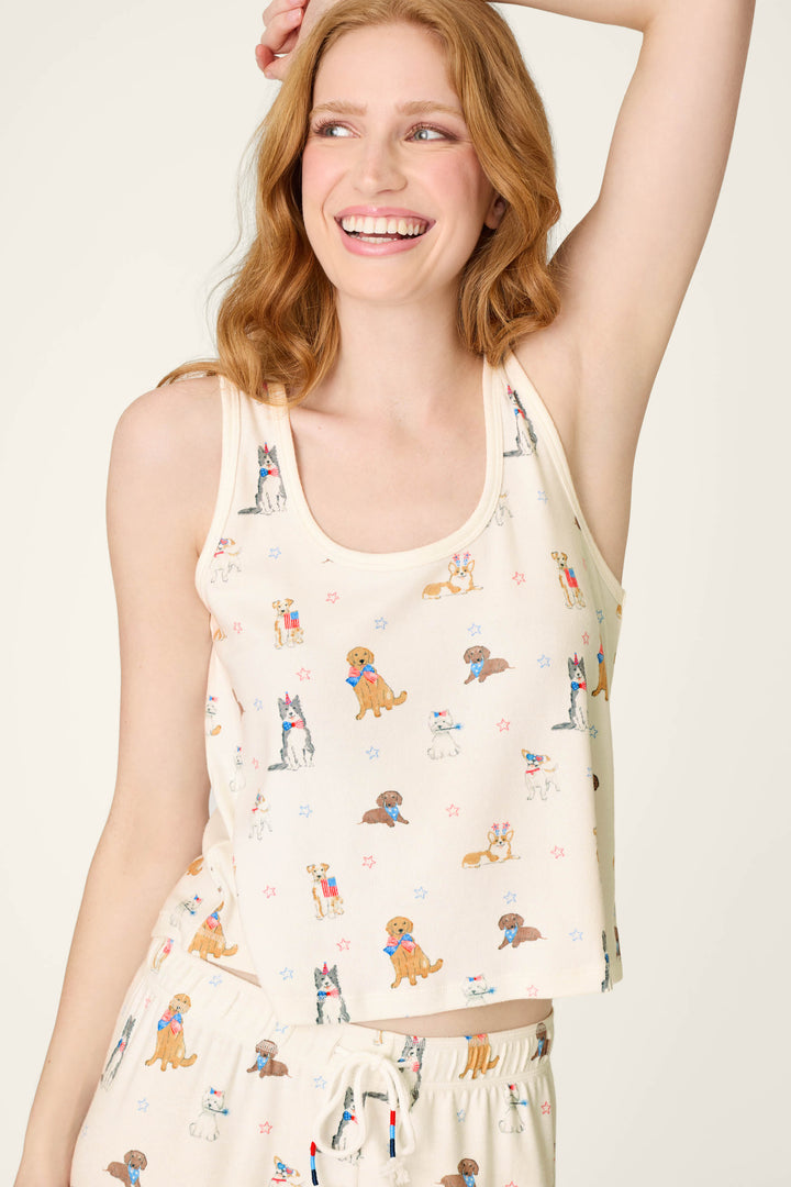 A smiling woman with red hair wears the P.J. Salvage Red, White, & Woof Patriotic Pups Tank Top, a cream scoop neck featuring a playful dog print. She poses with one arm raised, looking to the side against a neutral background.