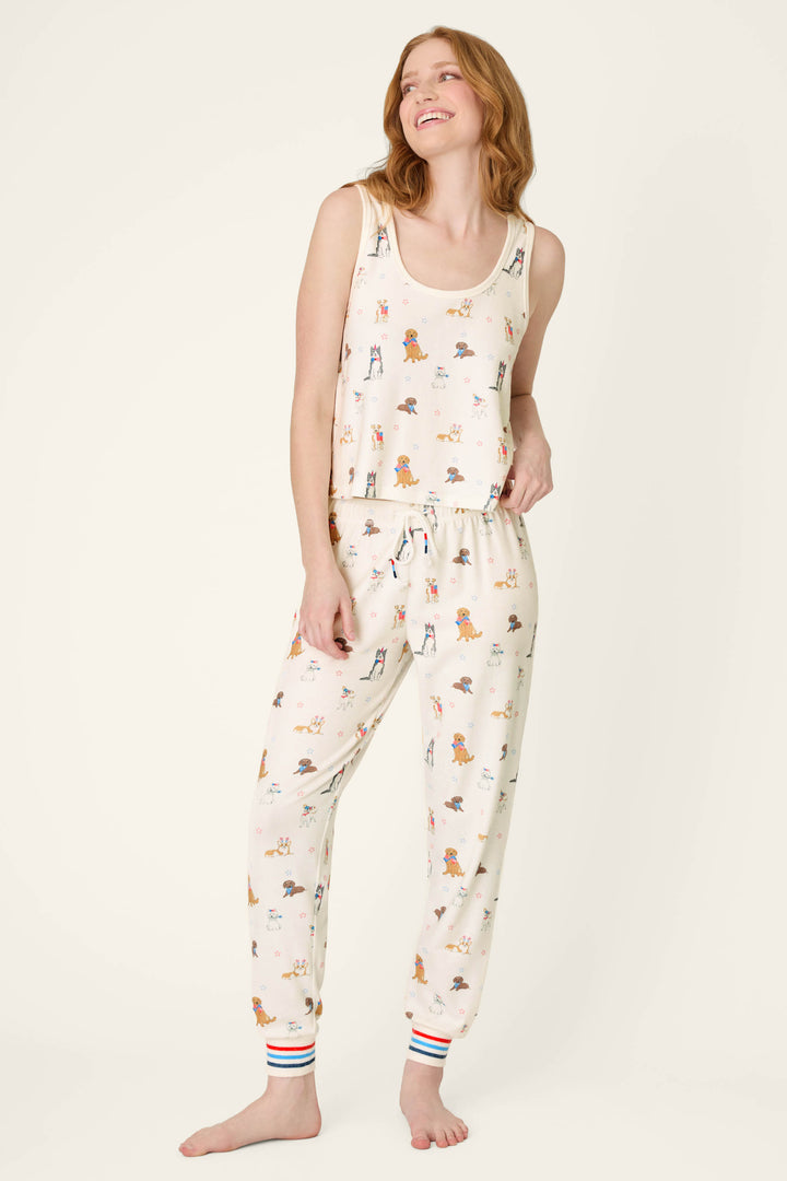 A smiling woman with wavy hair wears the P.J. Salvage Red, White, & Woof Patriotic Pups Tank Top—cream-colored pajamas featuring playful dog prints and colorful striped cuffs—standing barefoot against a plain background.