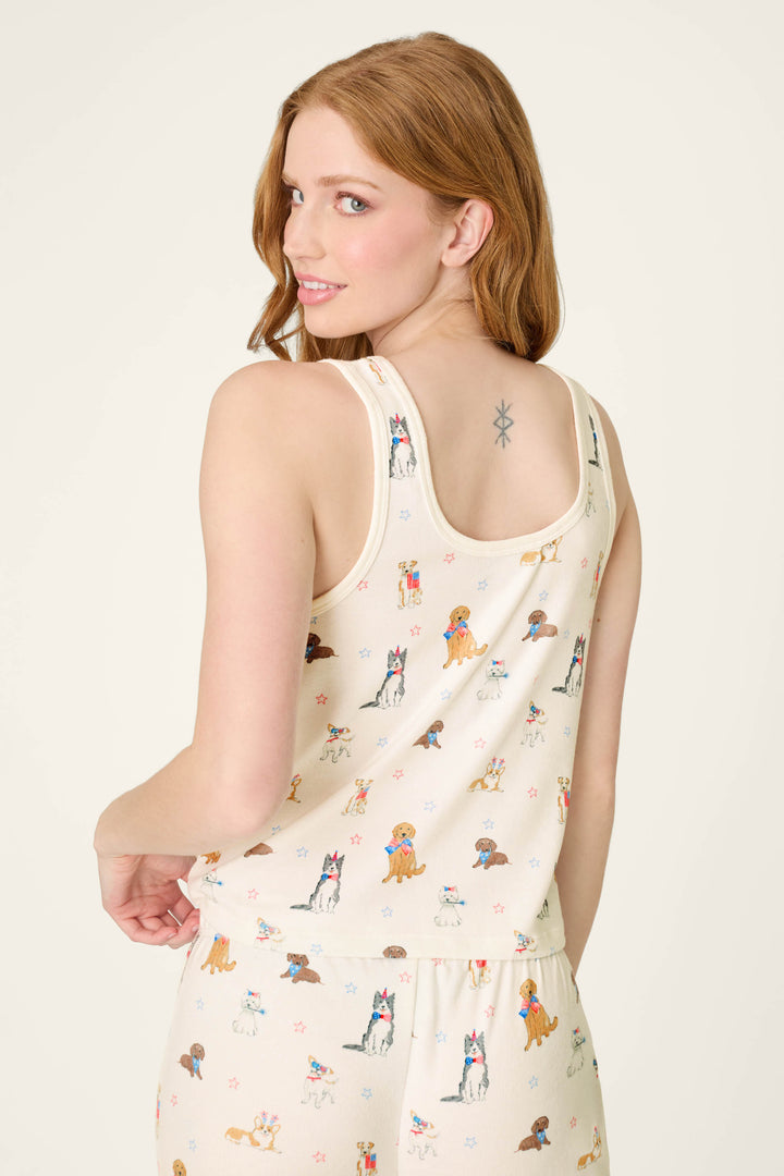A woman with long, light brown hair smiles over her shoulder, revealing a small star tattoo and wearing the P.J. Salvage Red, White, & Woof Patriotic Pups Tank Top in cream.